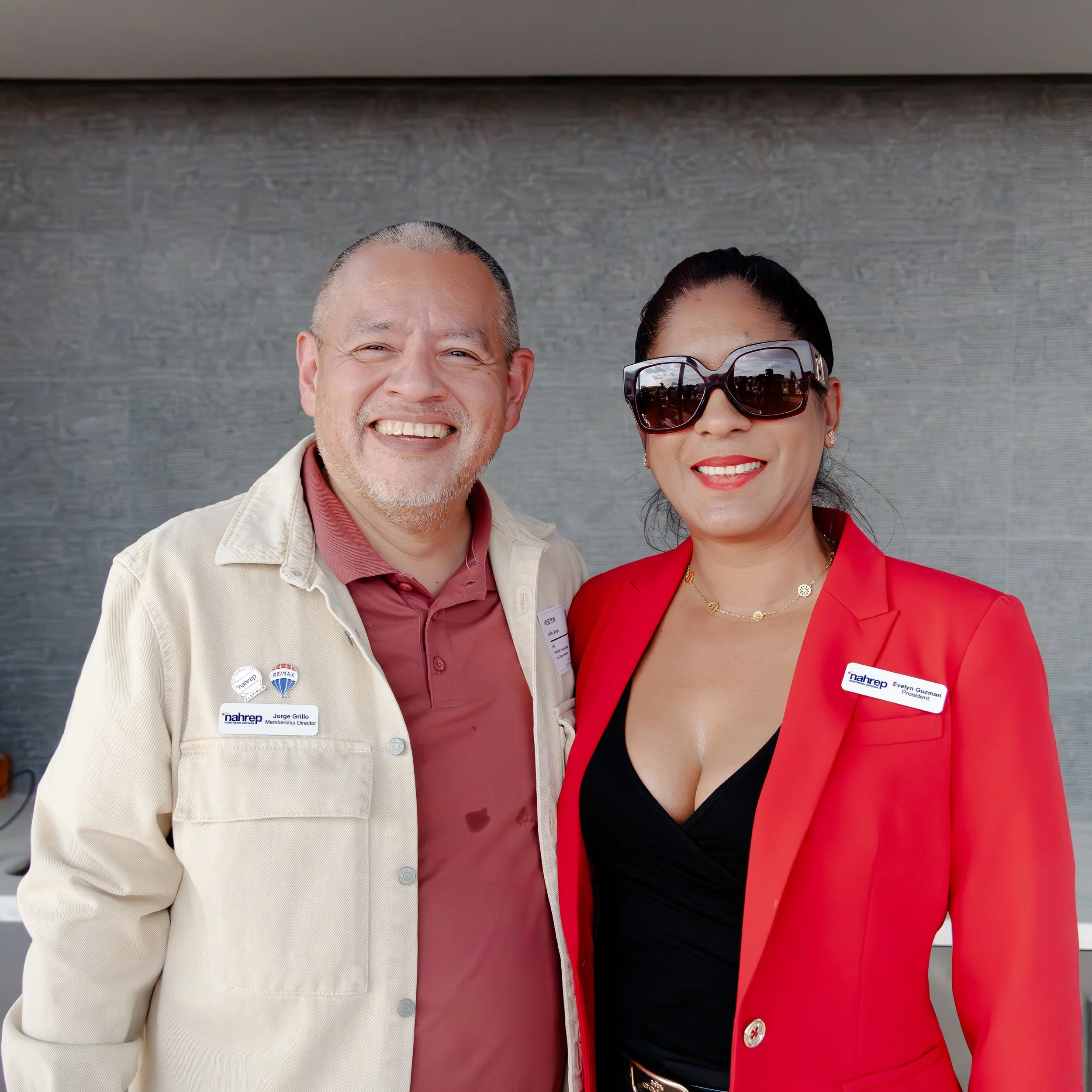 A man and woman posing together, smiling at the camera, indoors with a gray wall background. The man is wearing a beige jacket with badges, a maroon shirt, and has short gray hair. The woman is wearing large sunglasses, a red blazer, a black top, a g