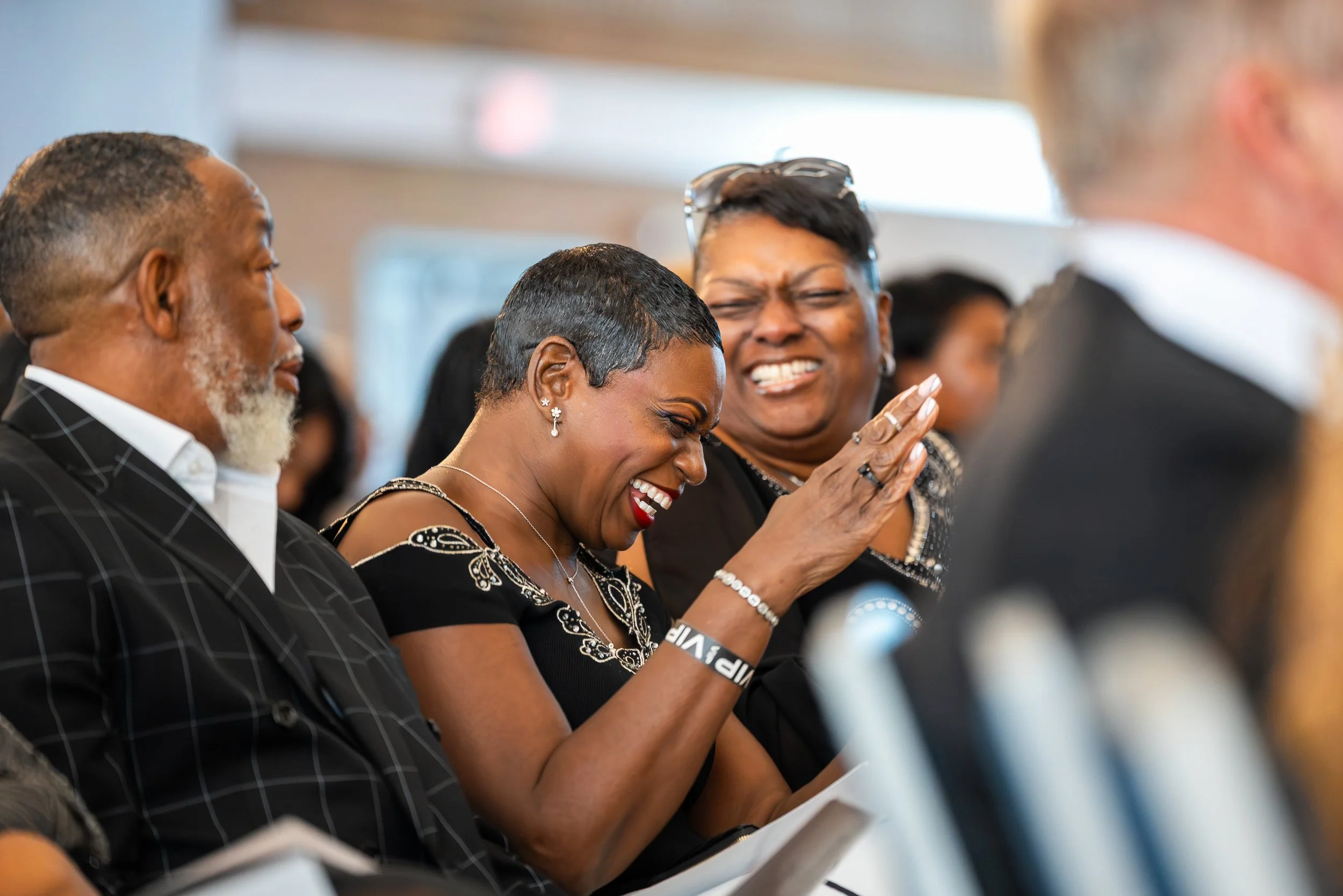 Three people sitting side by side, laughing and smiling during an event. The woman in the middle is laughing with her eyes closed, hand raised, wearing earrings and a black dress. The woman behind her has short hair, glasses on her head, and is also 