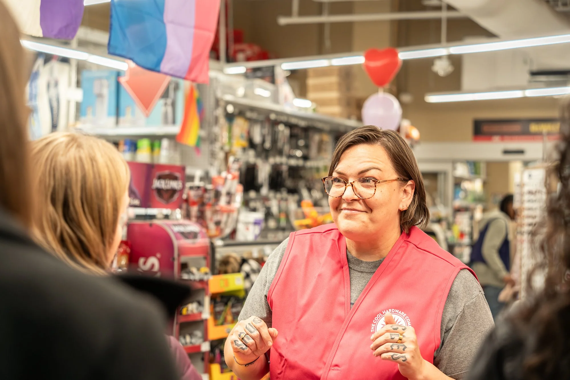 A woman with glasses and tattoos on her fingers, wearing a pink vest, is talking to customers inside a store, with shelves of snacks, toys, and merchandise in the background.