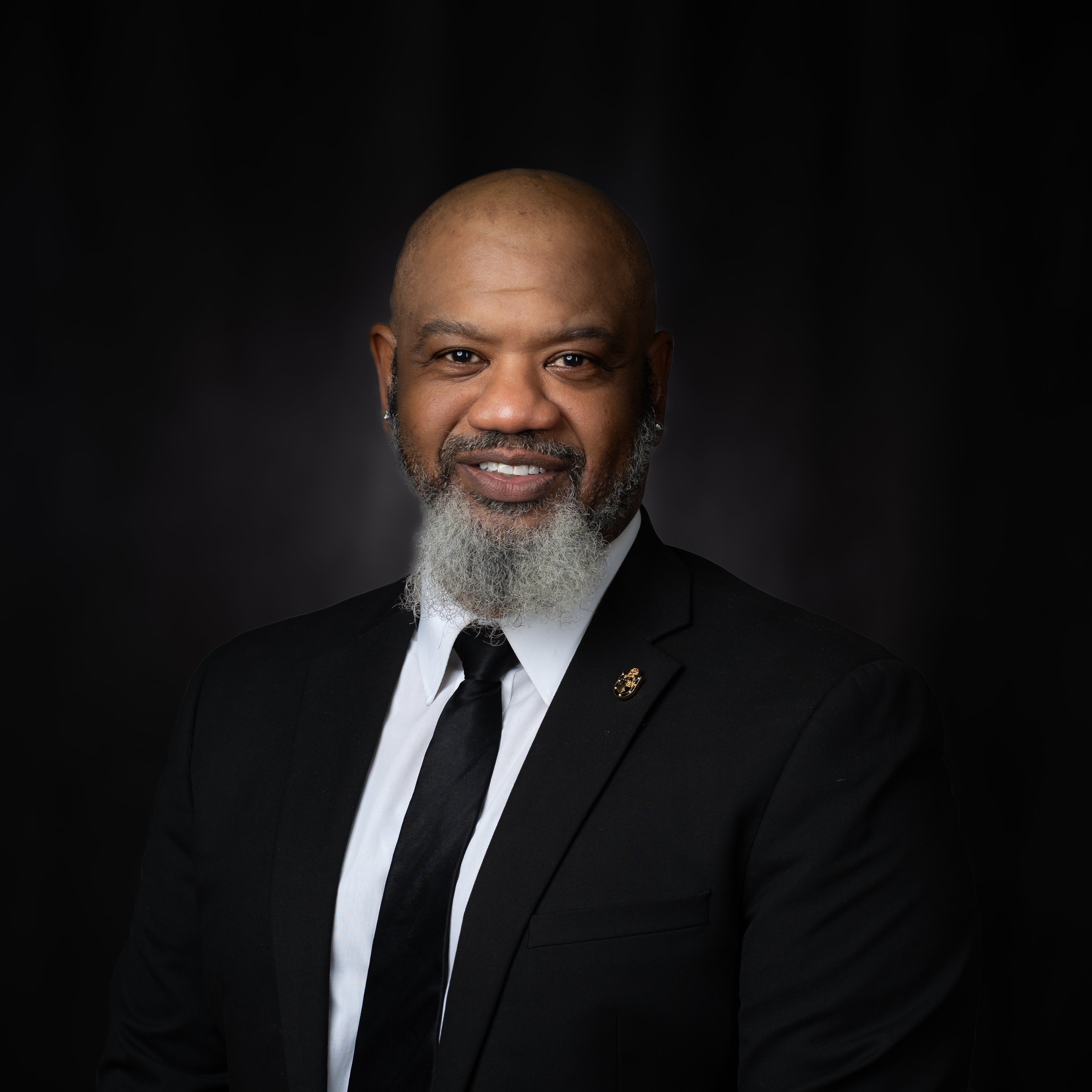Professional portrait of an African American man with a beard, wearing a black suit and tie, against a dark background.