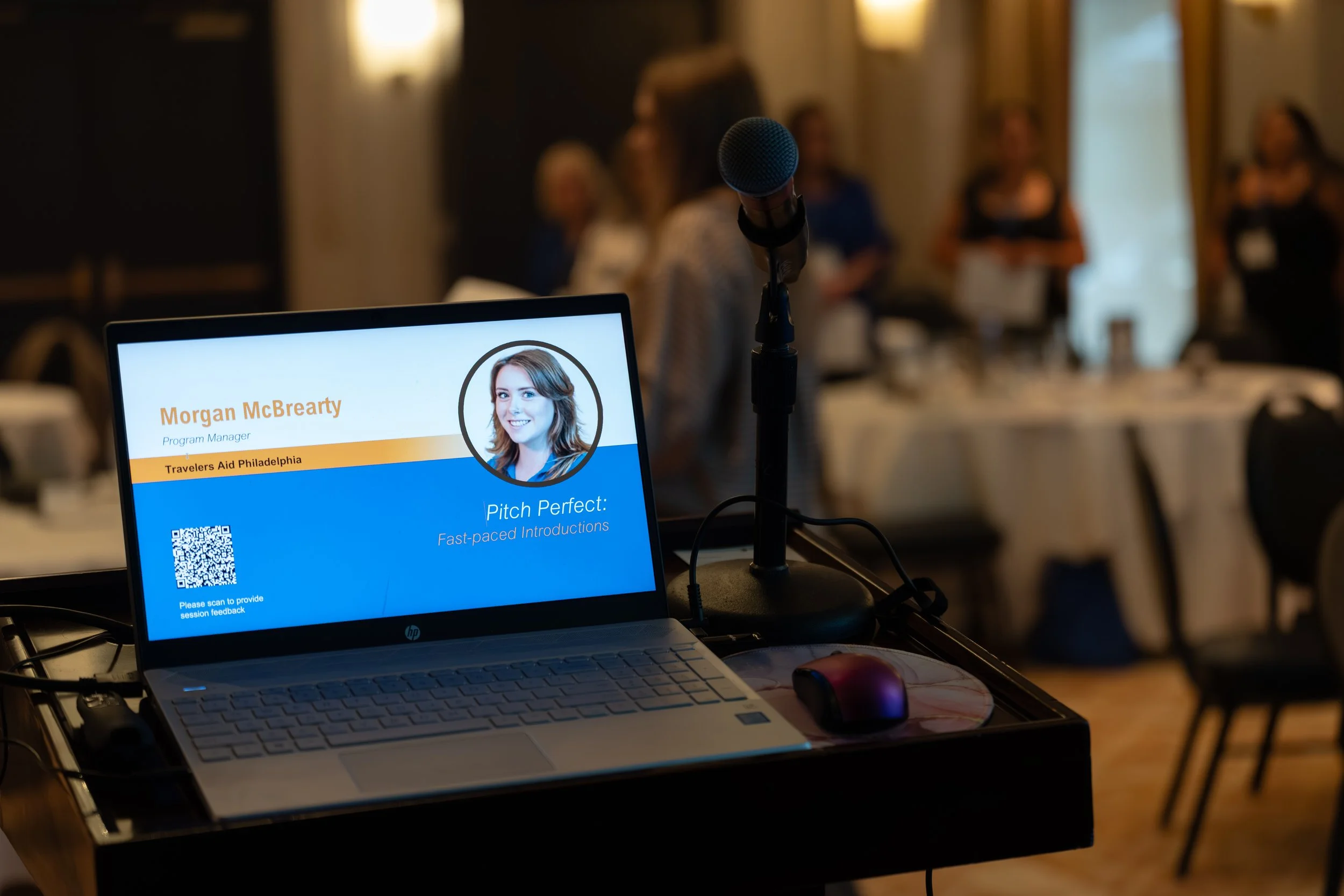 Laptop on a podium displaying a presentation slide with the name Morgan McBrearty, program manager for Travelers Aid Philadelphia, titled "Pitch Perfect: Fast-paced Introductions", with a QR code. A microphone is positioned next to the laptop. In the