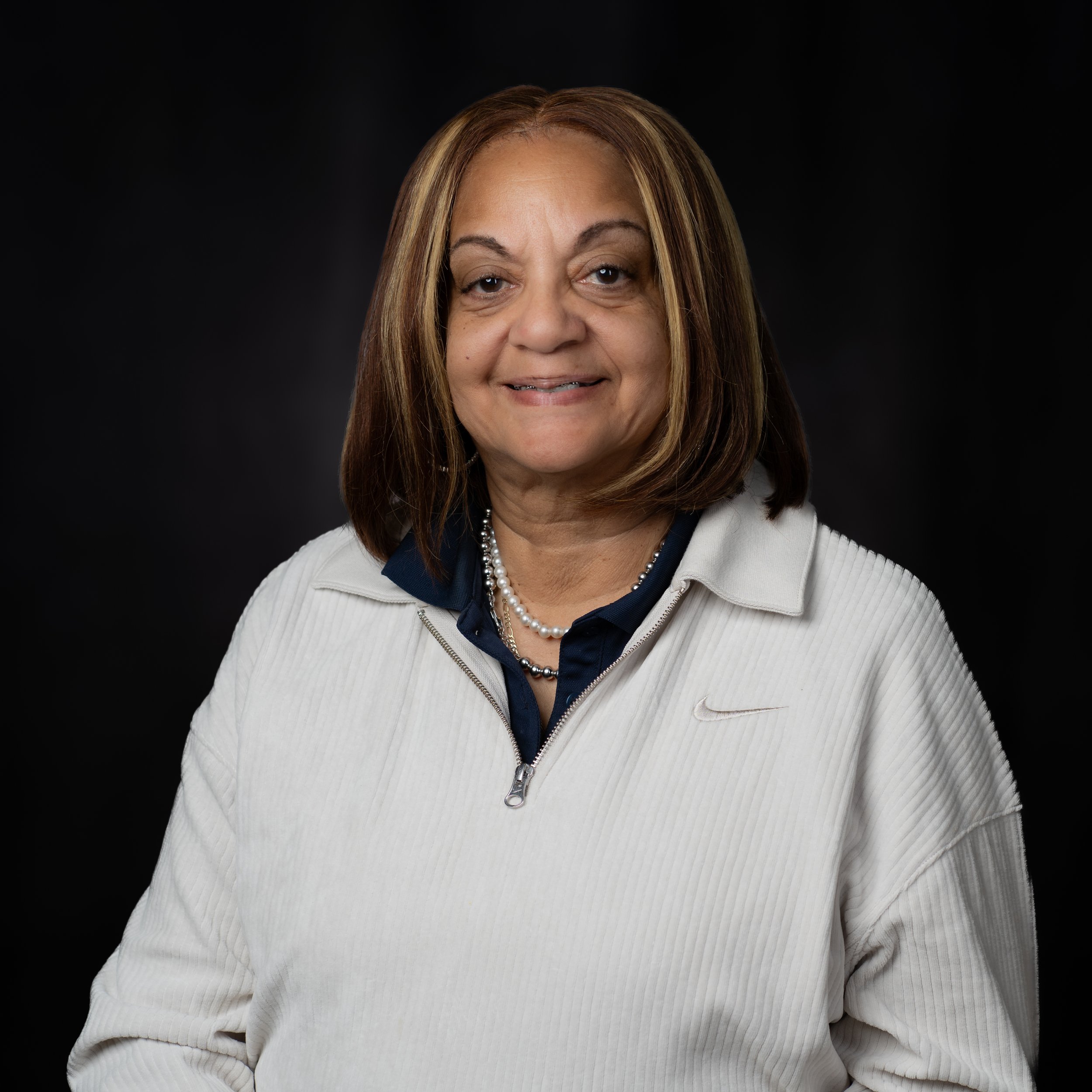 A middle-aged woman with shoulder-length brown hair. She is smiling slightly and is wearing a white Nike jacket over a dark-colored shirt, with pearl necklaces. The background is black.