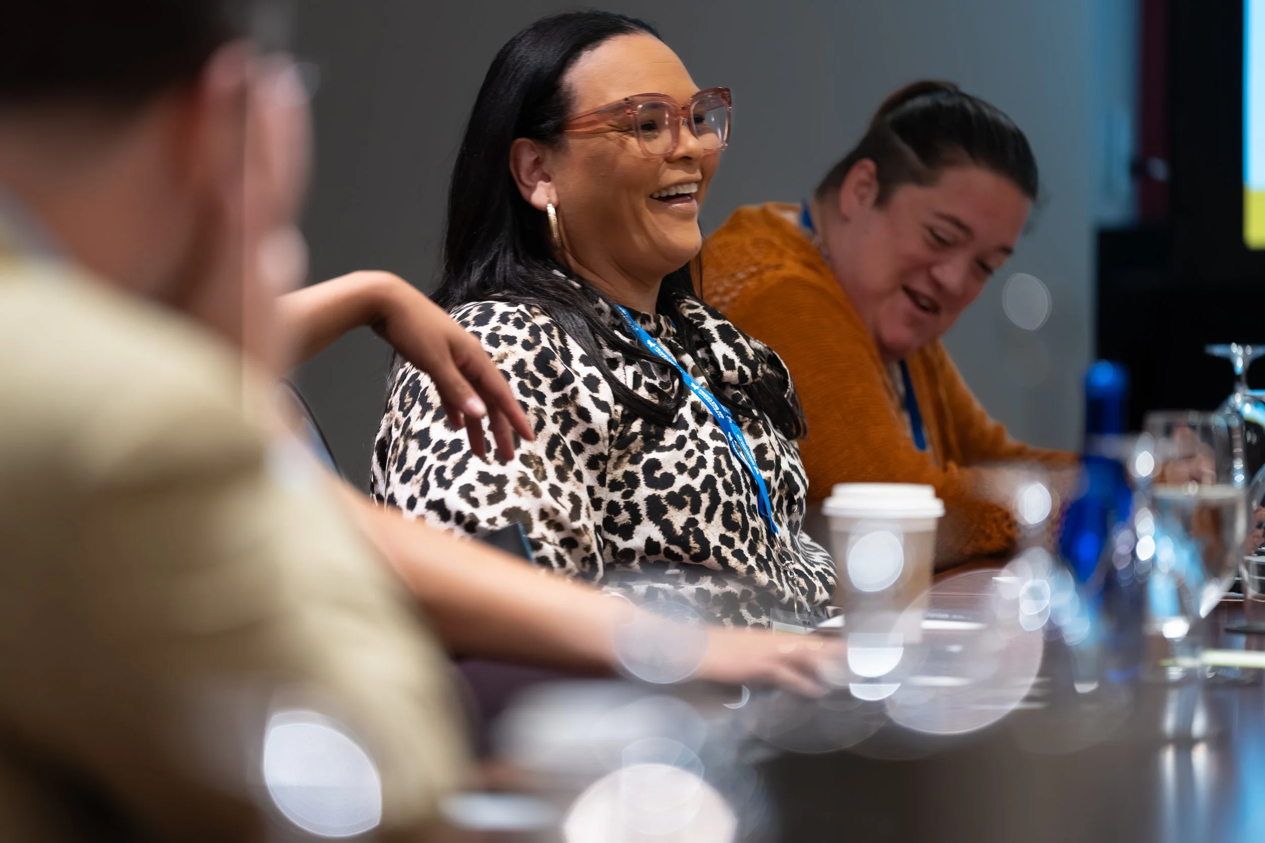 A woman with long dark hair, wearing large pink glasses and a leopard print top, smiling during a discussion at a conference table with other people.