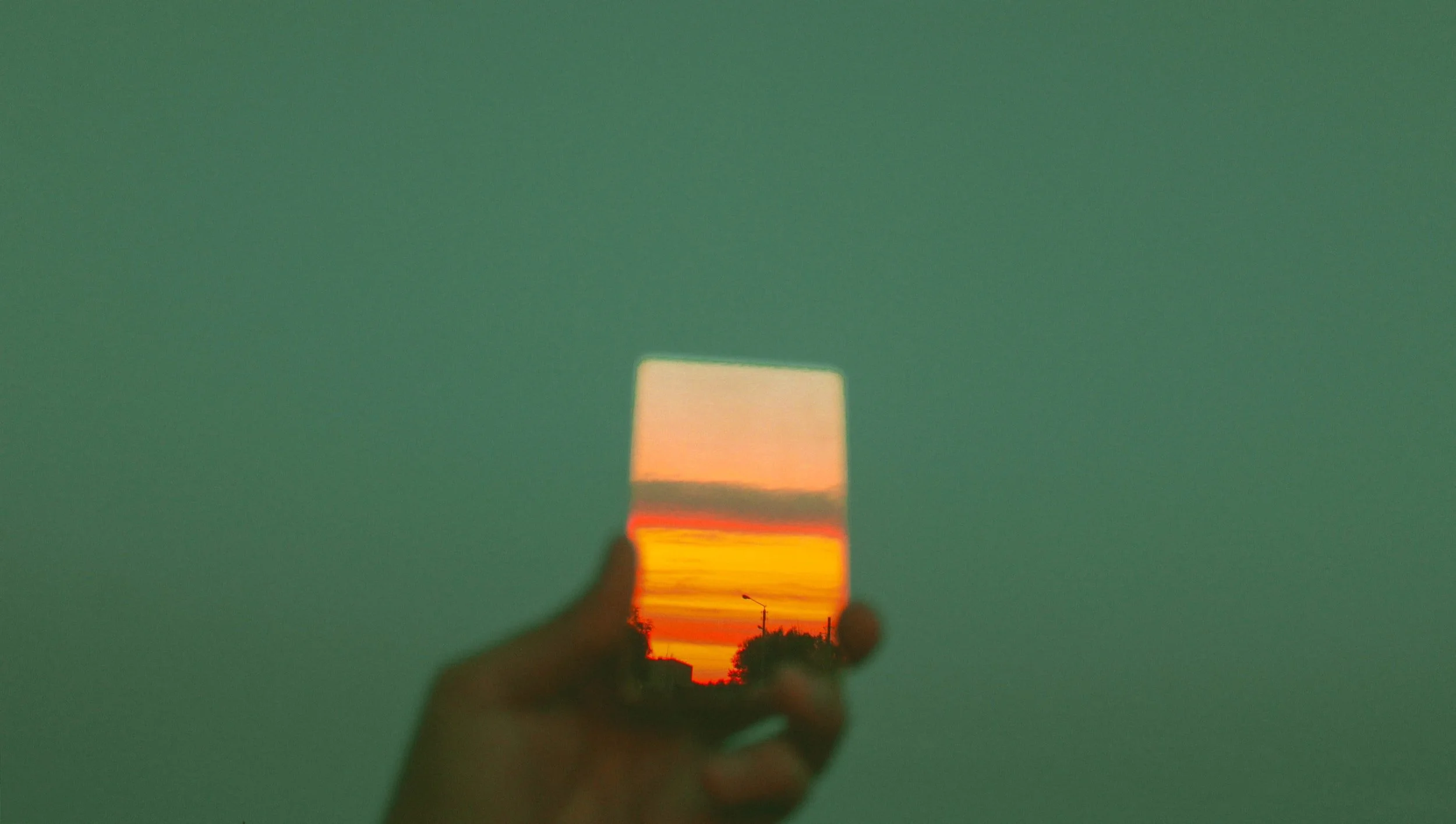 Person holding a mirror reflecting a sunset sky with orange, yellow, and pink clouds.