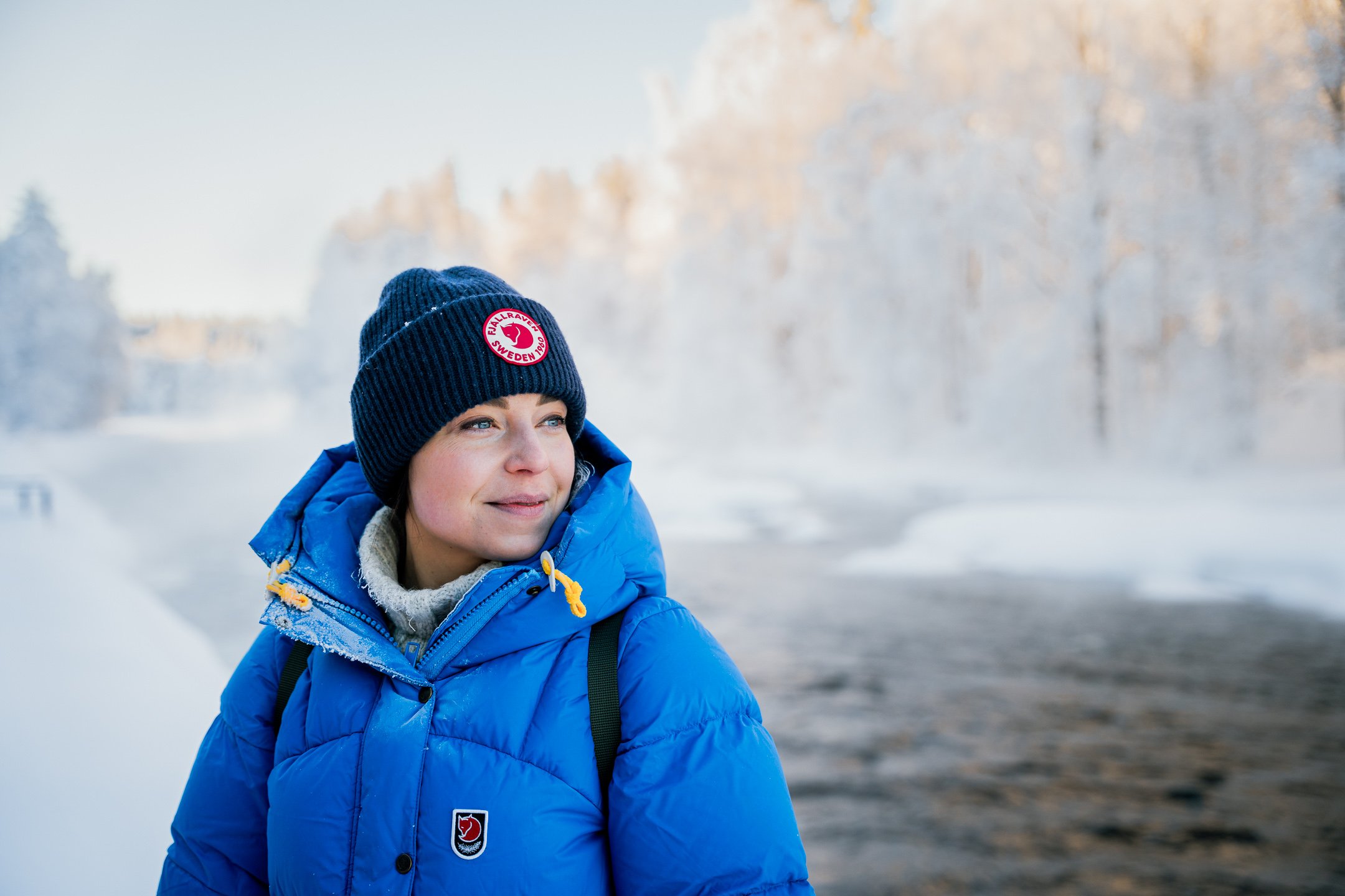 Woman wearing Fjällräven winter jacket and a beanie smiling and gazing towards the horizon next to frozen rapids during winter time in Kuhmo©Sami Laitinen.jpg