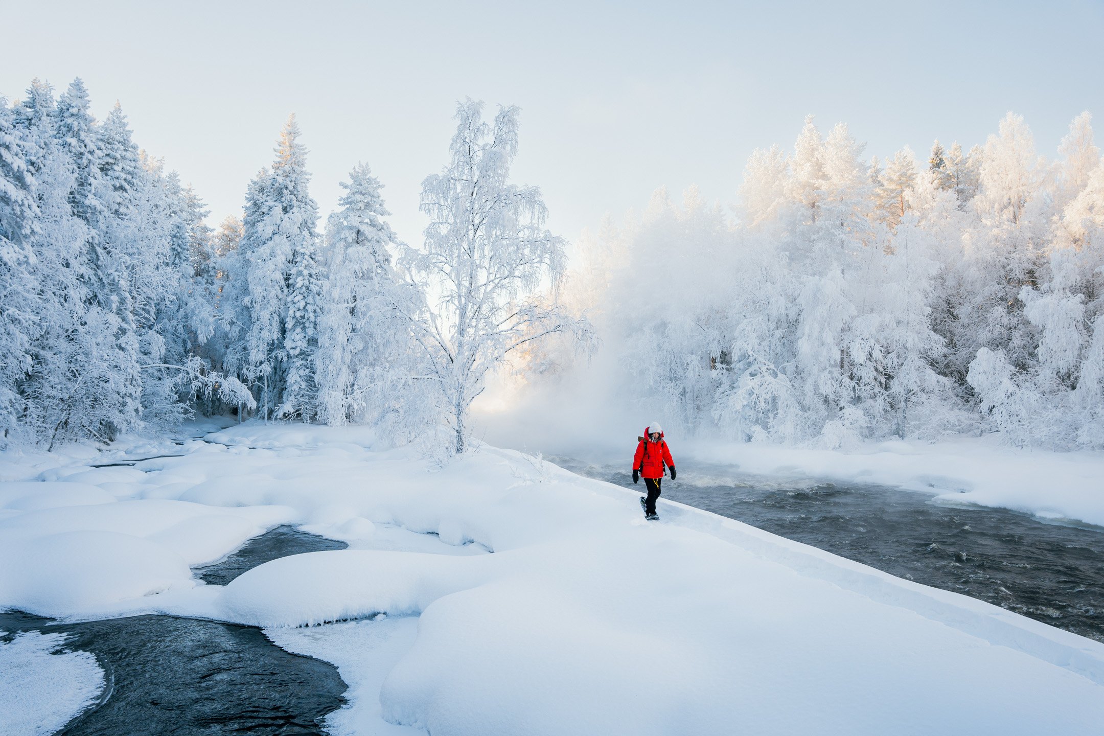 Person wearing red Fjällräven winter jacket walking next to rapids during a winter time in Kuhmo in Arctic Lakeland Kainuu©Sami Laitinen.jpg