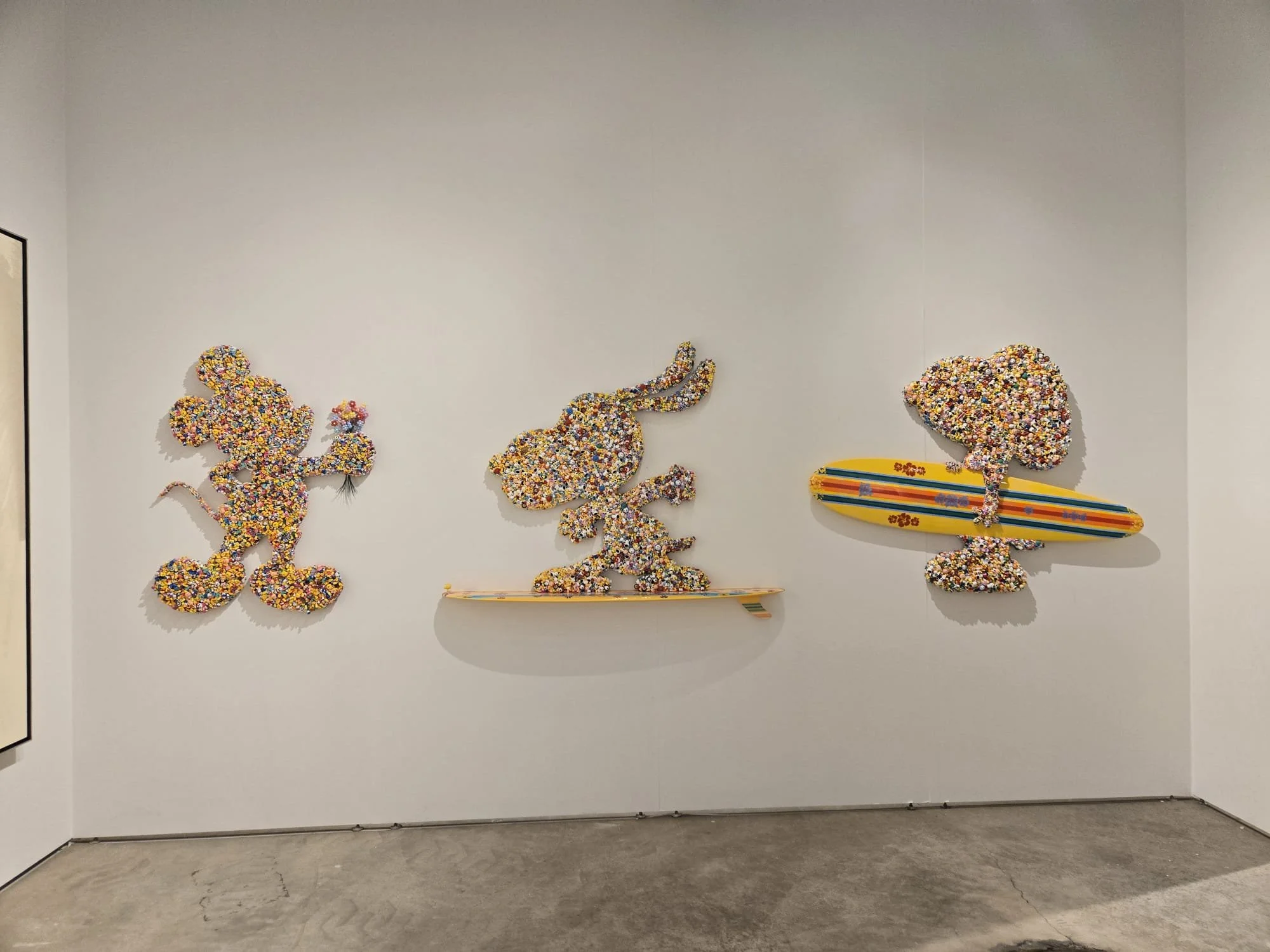 Three colorful mosaic art pieces of Mickey Mouse, a rabbit, and a surfboard with another Mickey Mouse, displayed on a white gallery wall.