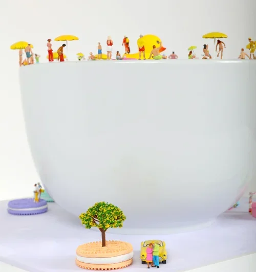Miniature figurines of people, umbrellas, and animals on top of a large white bowl, with small trees and cars at the base, creating a scene resembling a park or beach.