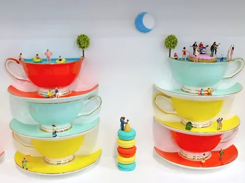 Stack of colorful teacups with tiny figurines of people and children placed on and around them, with a small moon decoration on the wall.