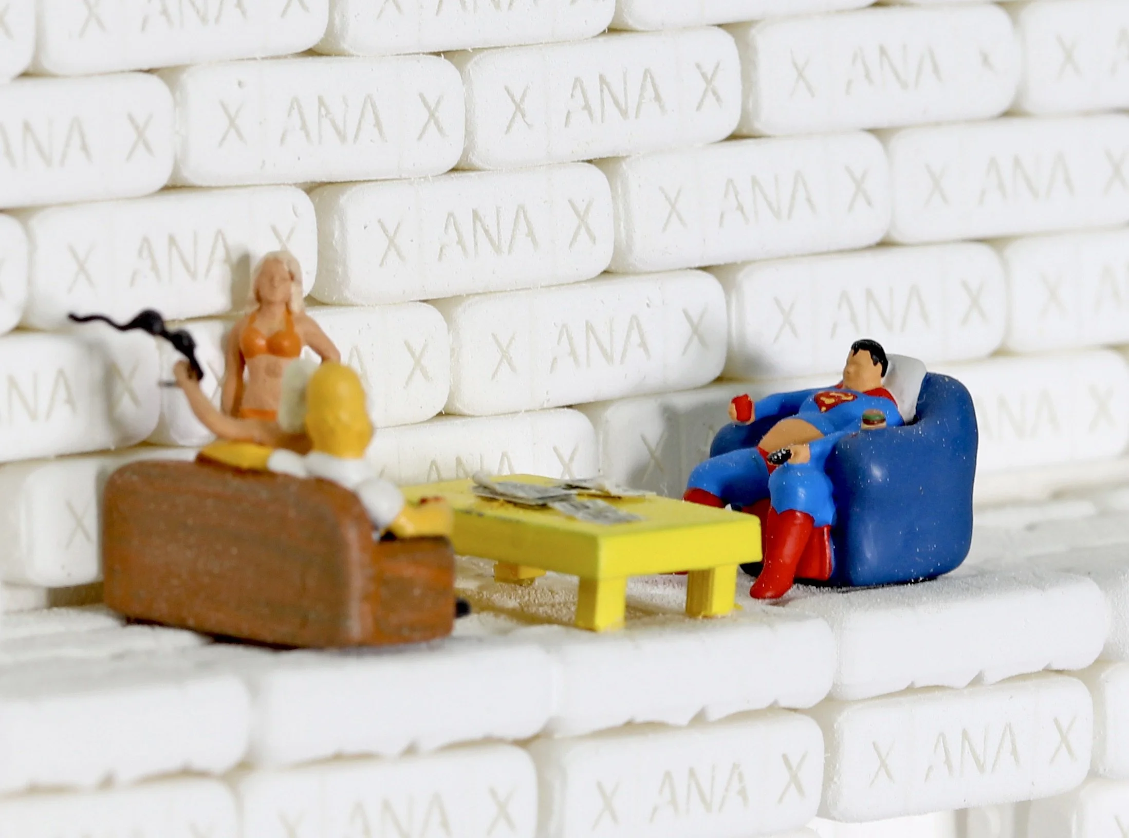 Miniature figurines of a woman sitting on a brown bench and a man lounging in a blue chair, watching TV, with a small yellow table between them, in front of a wall of white aspirin tablets labeled 'ANA'.