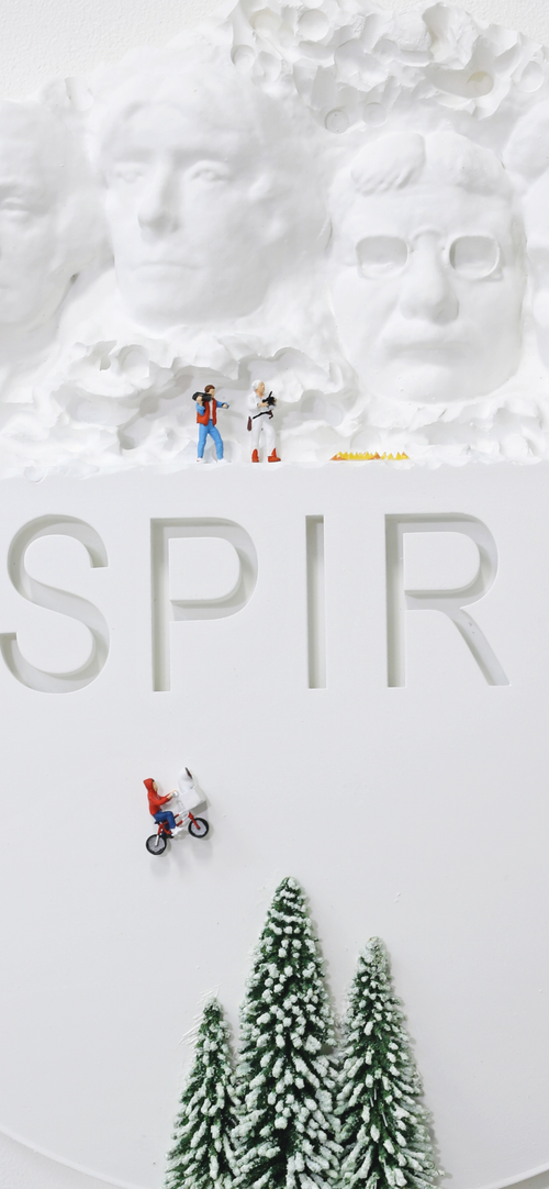 Miniature figures of two hikers with backpacks near Mount Rushmore, featuring large carved faces of U.S. Presidents, with the word 'SPIR' in large cut-out letters, a figure riding a bicycle, and snow-covered pine trees at the bottom.