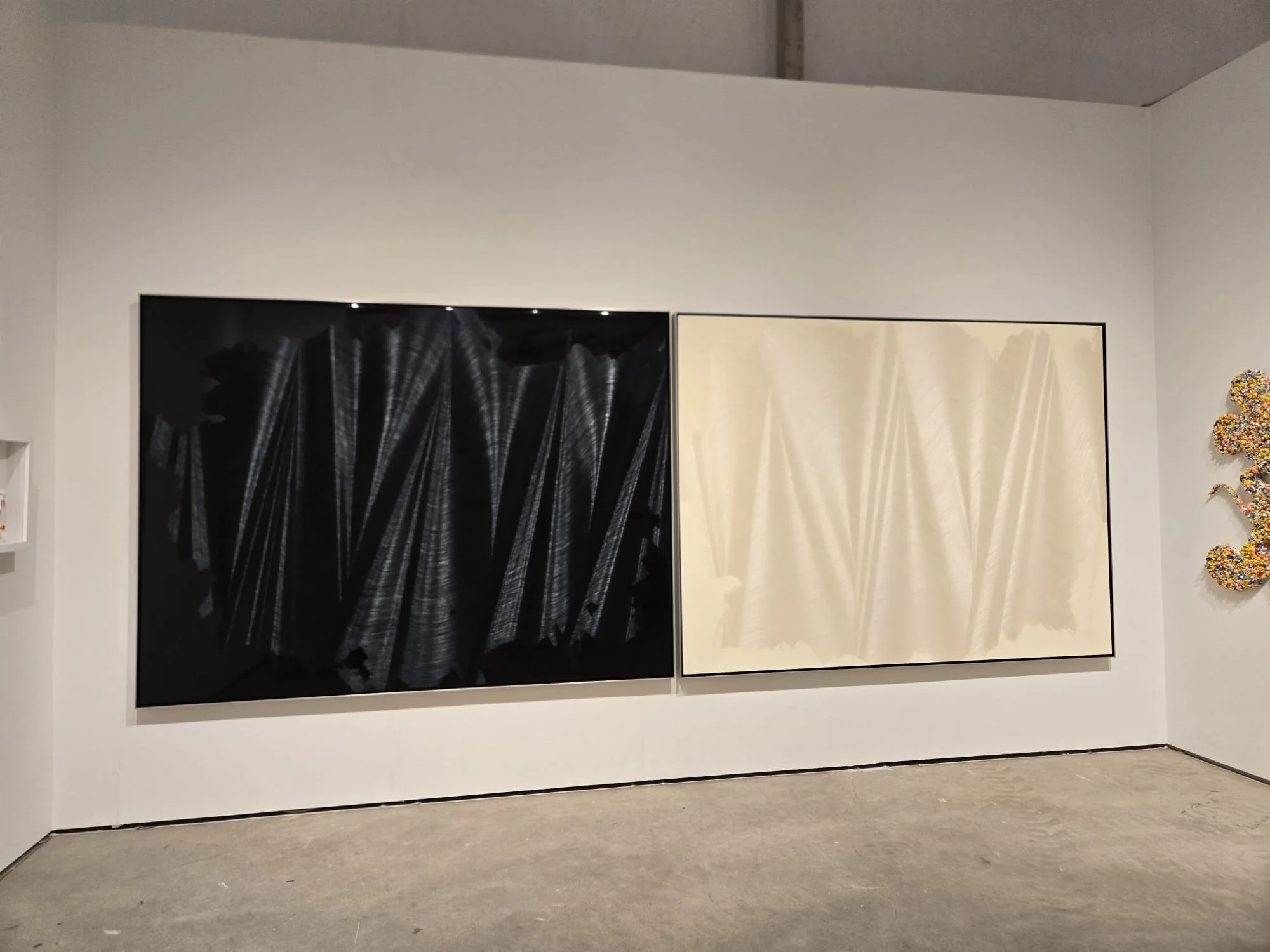 Two large abstract paintings on a white gallery wall, with the left painting in black and white and the right in shades of cream and beige.