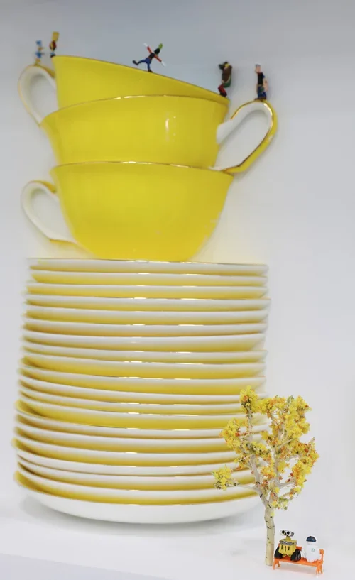 Stack of yellow teacups and white saucers with small toy figures and a toy tractor at the bottom right.