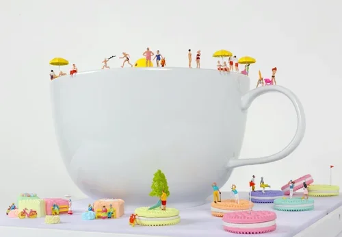 Tiny toy people standing and sitting on the rim of a large white teacup surrounded by colorful miniature macarons and desserts.