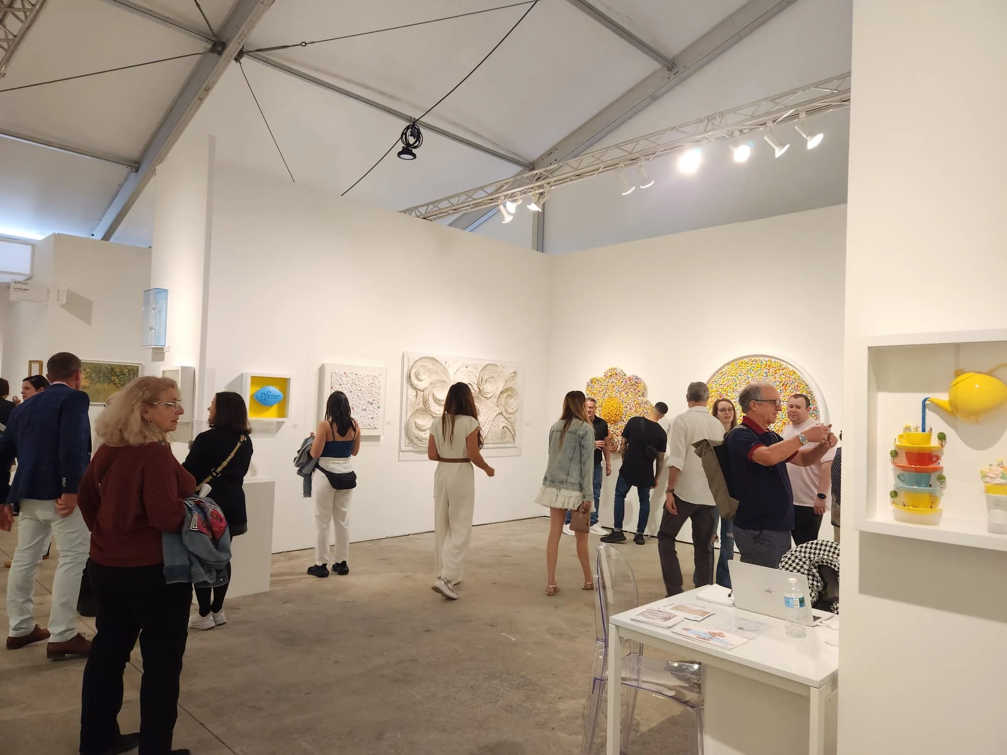 People viewing various artworks in an art gallery or exhibition space with white walls and ceiling, spotlights, and a few artworks visible on the walls.