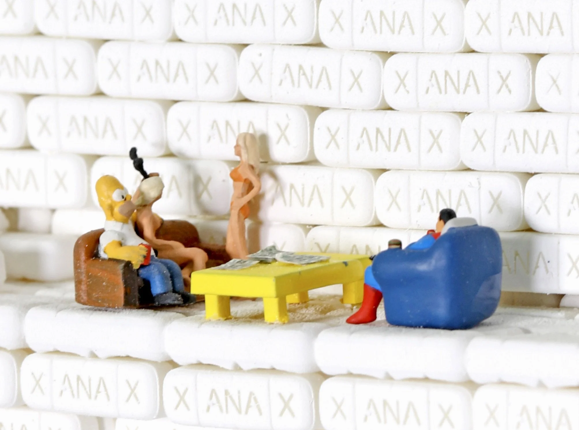Miniature figurines of characters from The Simpsons sitting around a small table, with a backdrop of white tablets with 'ANA' and 'X' printed on them.