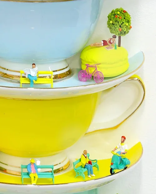 Colorful stacked teacups with miniature figurines sitting or standing on the edges, including a person on a yellow bench, a person on a teal bench, a person riding a scooter, and a person reading a book under a tree with a pink bicycle nearby.