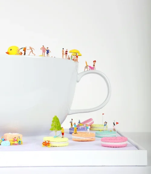 Miniature people and animals on the rim of a large white teacup, with small, colorful macarons and toys below.