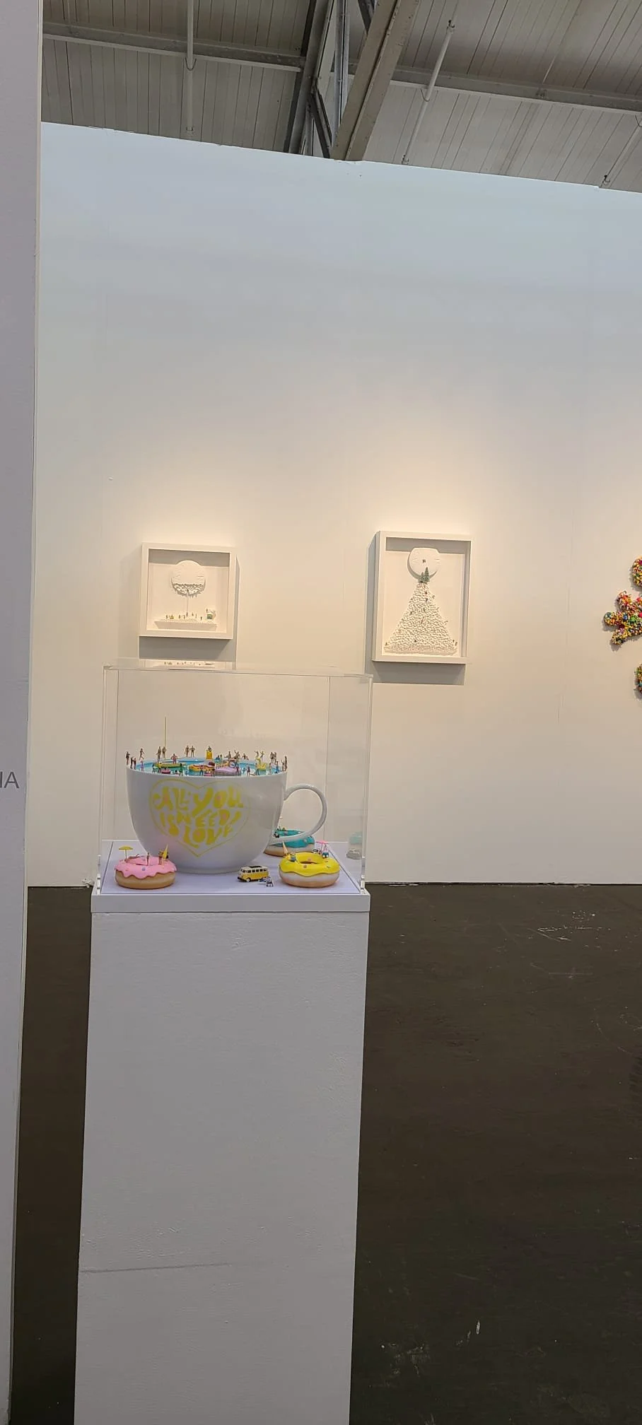 Art display in an art gallery featuring miniature figurines inside a large coffee cup and two decorated cookies on a white pedestal. Artwork hangs on a white wall in the background.
