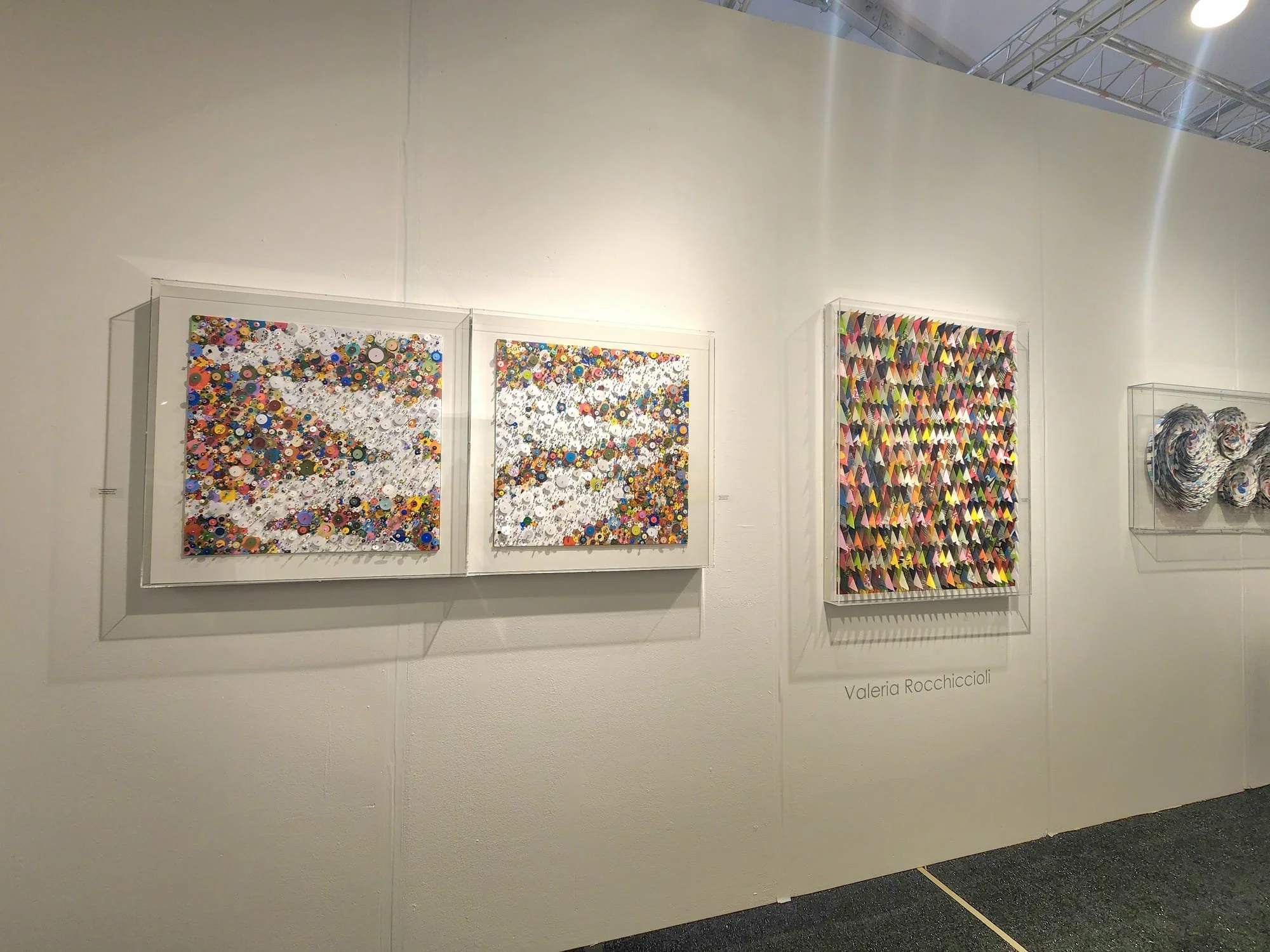 art exhibit featuring colorful mixed media artwork, including two square pieces with buttons, a larger rectangular piece with folded paper, and a sculpture resembling swirling paper, all displayed on a white gallery wall.