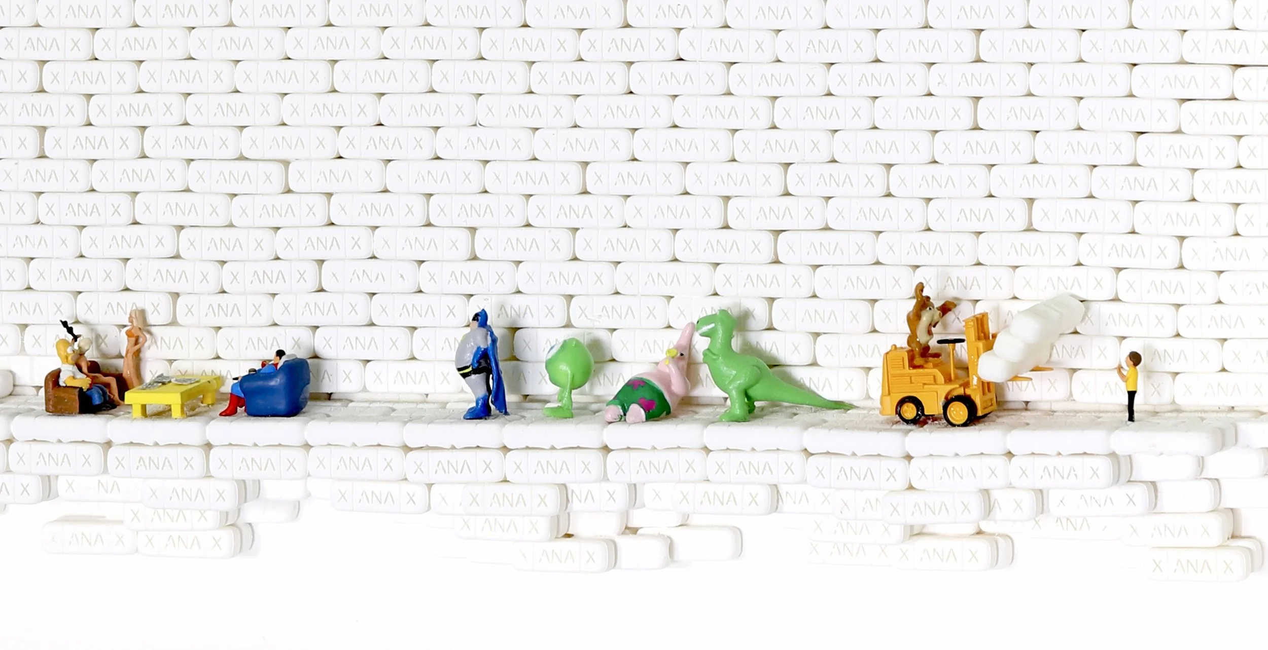 Miniature toy figures arranged on a surface in front of a wall made of white oval pills with 'ANA' inscribed on them, representing a creative scene.
