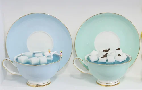 Two teacups with saucers, one light blue and one mint green, filled with ice and small items that resemble miniature food or decorations.