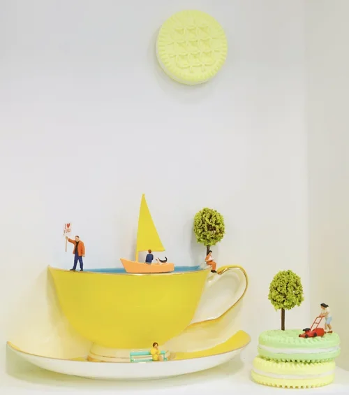 Decorative miniature scene with a yellow teacup and saucer, containing a small sailboat, miniature trees, and tiny figurines, with a large yellow cookie or pastry shaped object on the wall above.