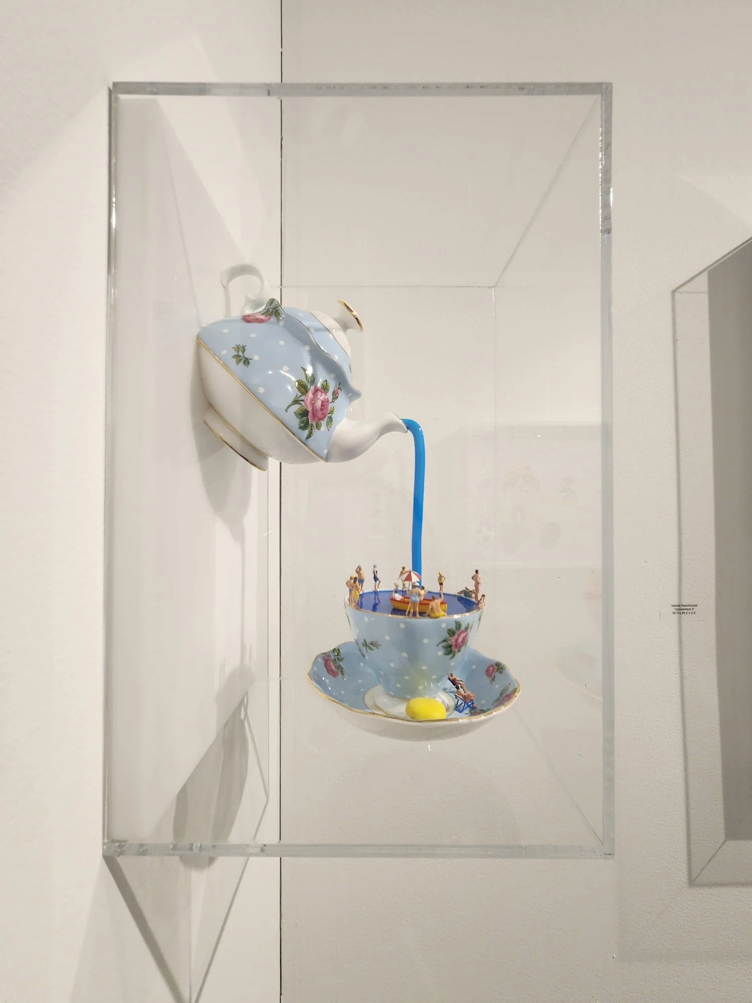 A teapot pouring water into a teacup with miniature figures of people and umbrellas, all displayed in a glass case