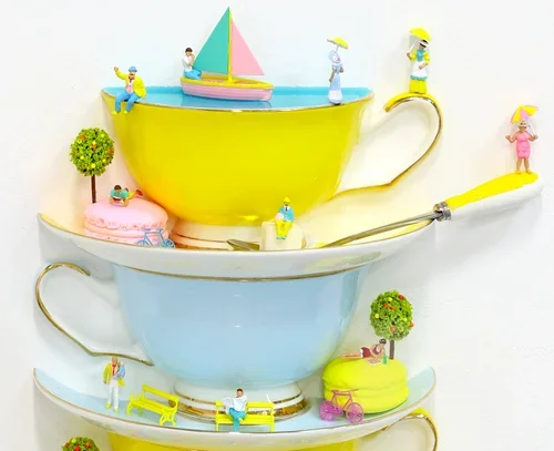 Stacked teacups with miniature figurines of people engaging in leisure activities, decorated with small trees, a pink cake, and a toy sailboat on top.