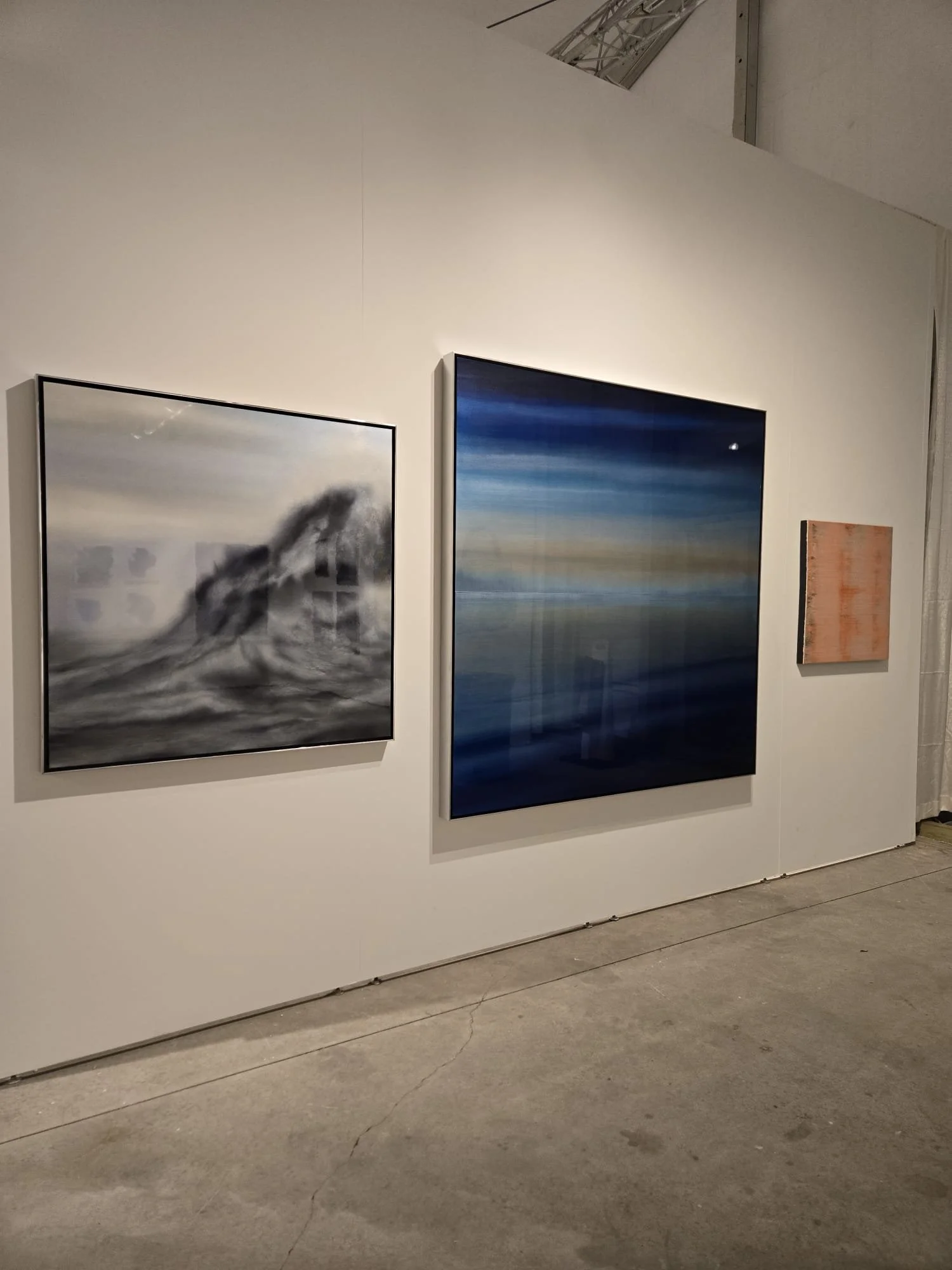 Three paintings on a white gallery wall, featuring abstract and landscape styles, with a concrete floor below.