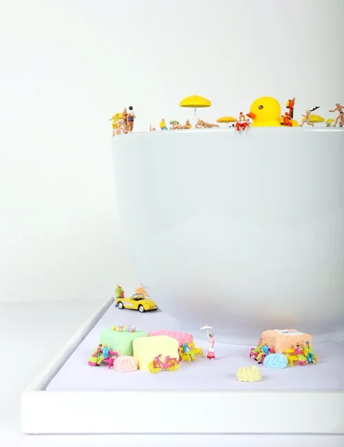 Miniature toy beach scene with small figures, umbrellas, and a rubber duck on a white surface.
