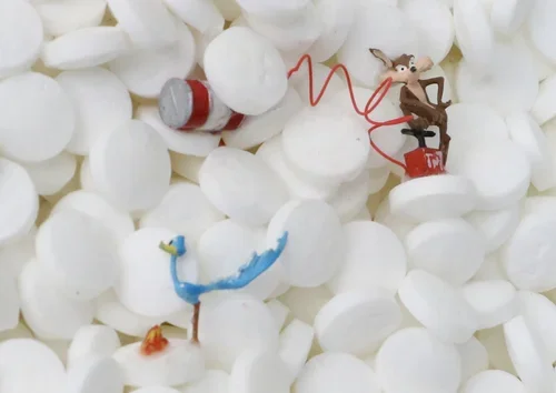 A small figurine of Jerry from Tom and Jerry is sitting on a red piece of cheese, holding a red wire with a zigzag pattern, surrounded by white packing peanuts.