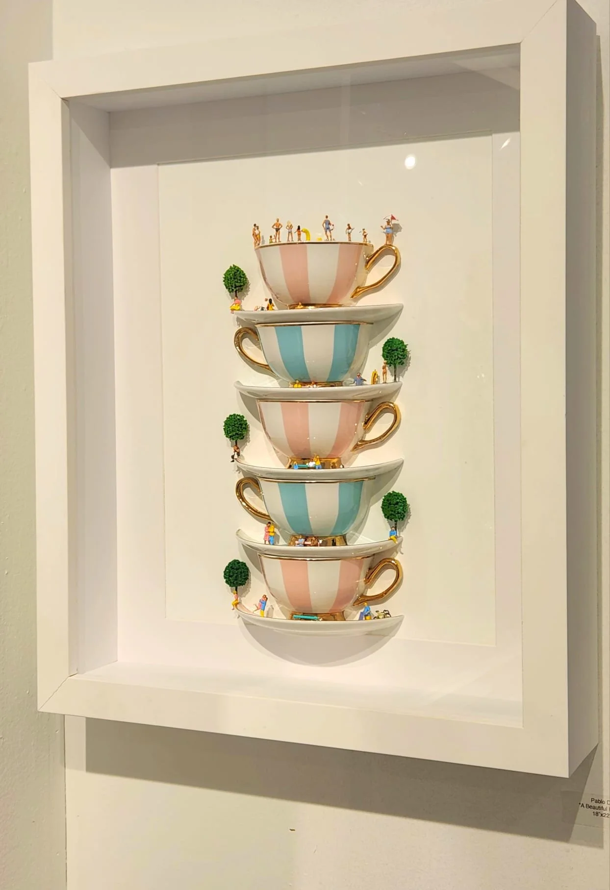 Decorative art piece featuring stacked teacups with miniature people and trees, displayed in a shadow box.