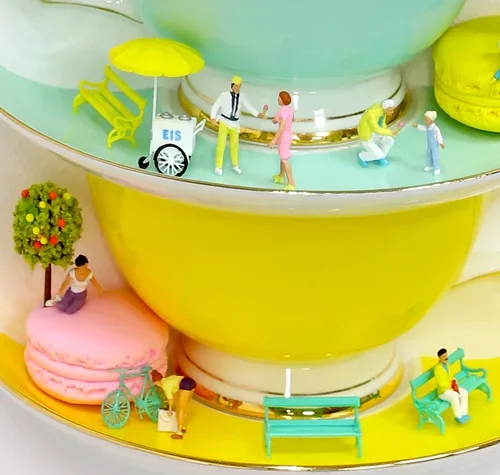 Miniature figurines of people near a yellow and green teacup, some sitting on benches, others walking, with a tree and an ice cream cart in the scene.