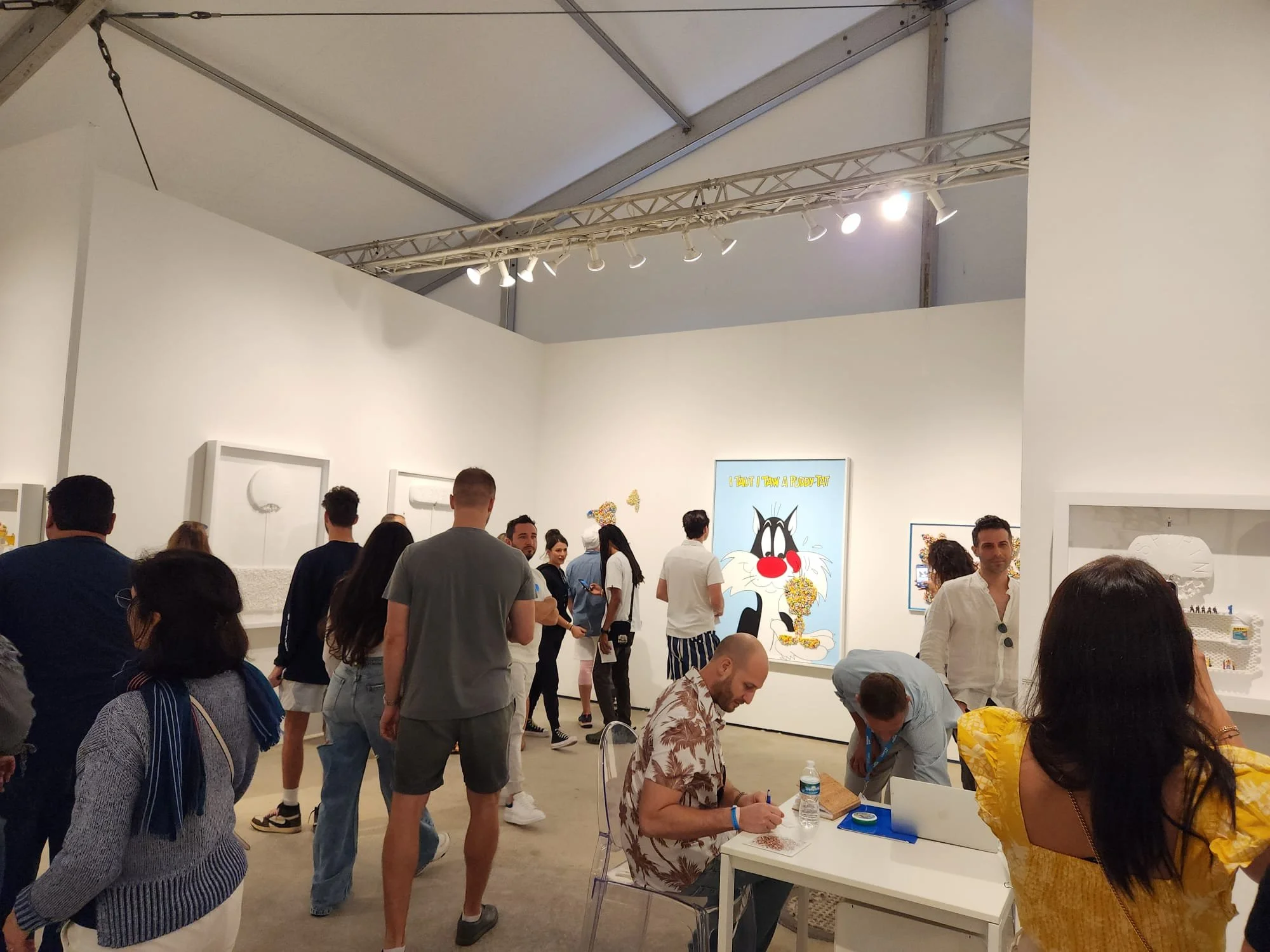 At an art gallery, people are viewing artworks and engaging with exhibits. The space has white walls, a high ceiling with spotlights, and various people of different ages and styles.