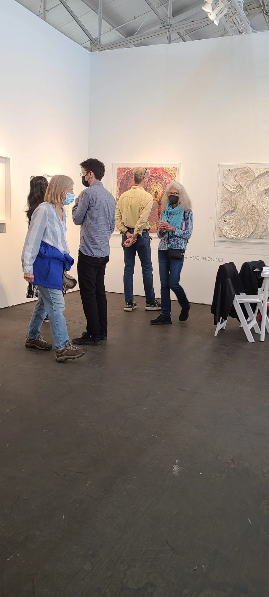 People wearing masks at an art gallery, looking at and discussing paintings on the wall.