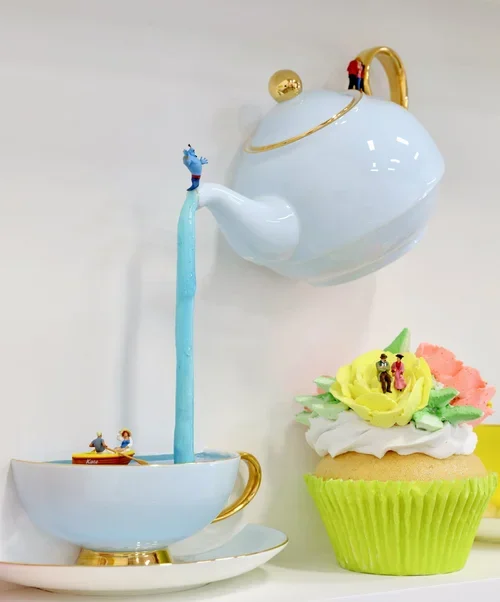 A miniature ceramic scene of a tea cup with a blue stream of water, a large teapot pouring water, and a cupcake with yellow frosting and small figurines on top.