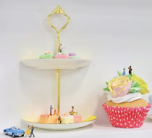 A two-tiered white and gold decorative stand with small figurines on and around it, placed next to a large decorated cupcake with made of colorful icing and tiny figures, on a white surface.