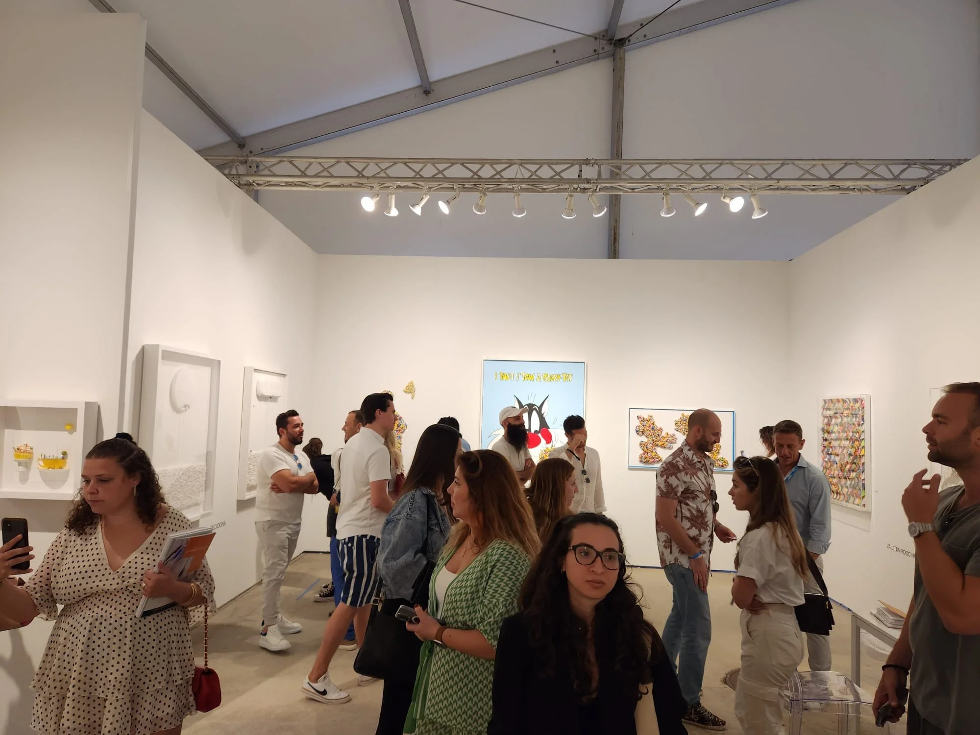 People attending an art gallery exhibition in a white-walled space with artwork displayed on the walls, some people are looking at the art, talking, or taking photos.