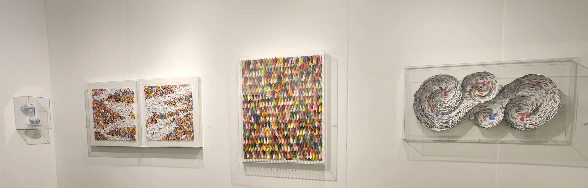 Art exhibit displays four mixed media artworks on a white wall, featuring colorful buttons, paper origami, and rolled paper sculptures encased in glass.