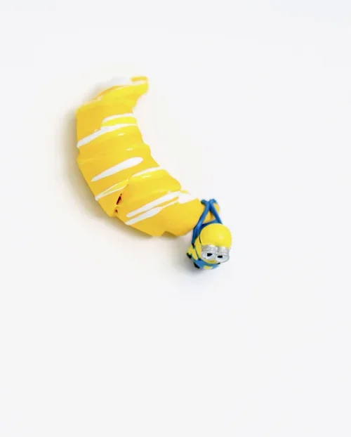 A Minion toy wrapped in a banana peel with white icing detail, lying on a white surface.