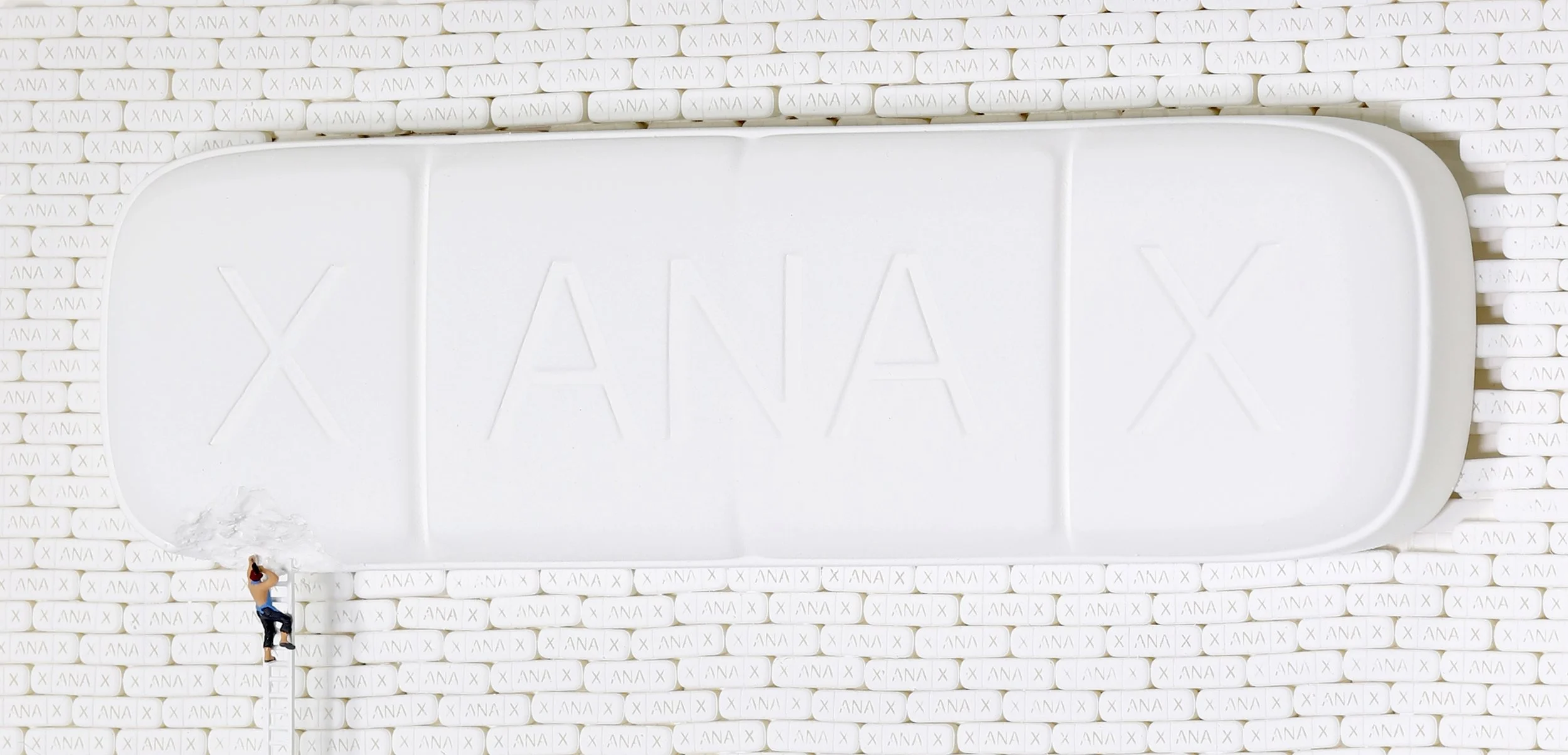 Close-up of a large white tablet with the brand name 'XANAX' embossed on its surface. The background consists of numerous white pills engraved with 'X' and 'ANA'.