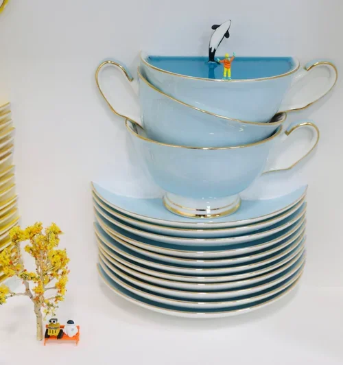 Stacked white teacups with gold trim on top of matching saucers, with a small toy penguin figurine and a bigger toy figure of a person in a yellow outfit on top of the top cup, and a small yellow figurine with a black hat near a small yellow tree on 