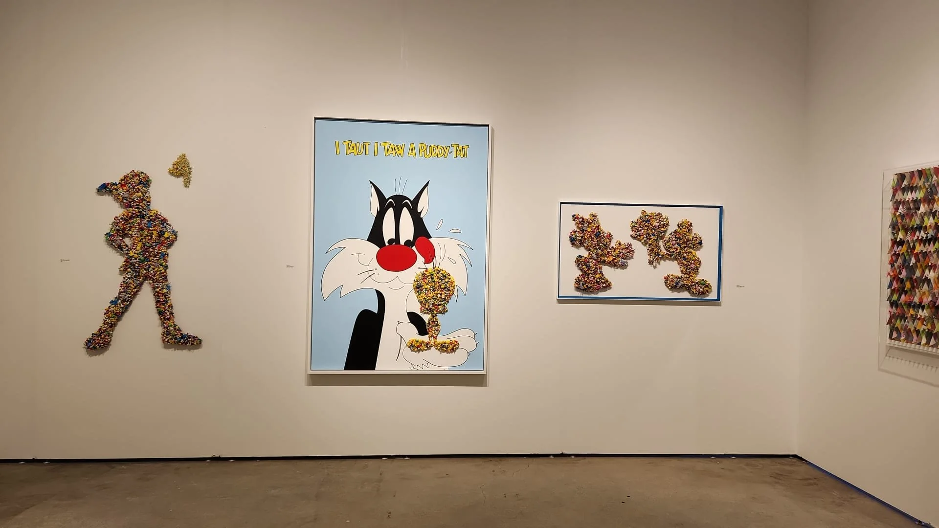 Art gallery wall showing various colorful pop art pieces including a large cartoon of a black and white cat licking a multicolored ice cream cone, with the words "I TAUT I TAW A PUDDY TAT" above, a silhouette of a woman made of small, colorful object