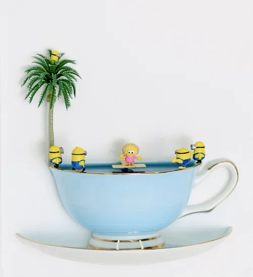 A light blue teacup with a matching saucer, decorated with small yellow Minions surrounded by a tiny figure of a girl, a palm tree, and a yellow minion figurine on top.