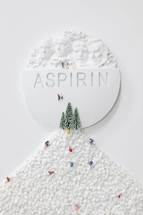 An artistic display of the word 'ASPIRIN' on a white circular background with carved faces at the top, snow-covered trees, and miniature figurines skiing down a mound of white pills.