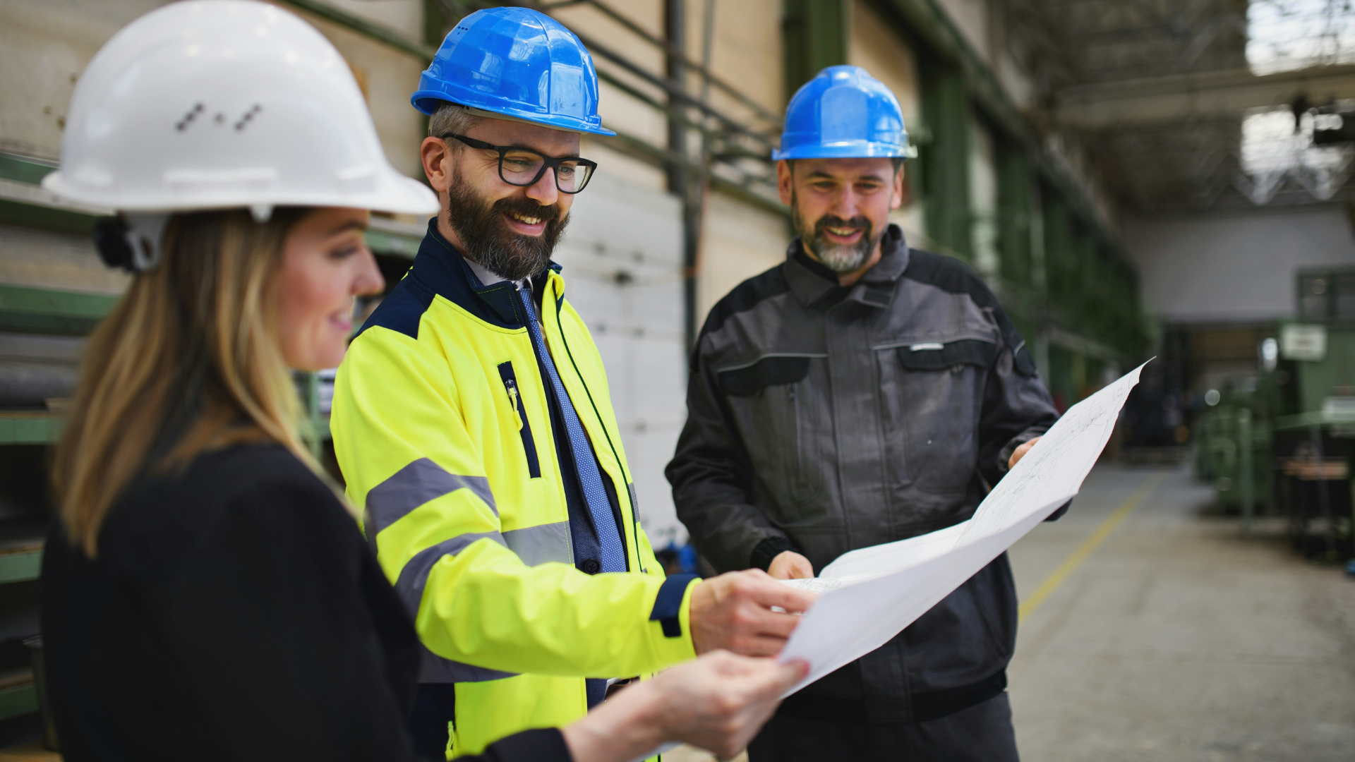 Three construction workers in safety helmets and apparel discussing plans inside an industrial warehouse.