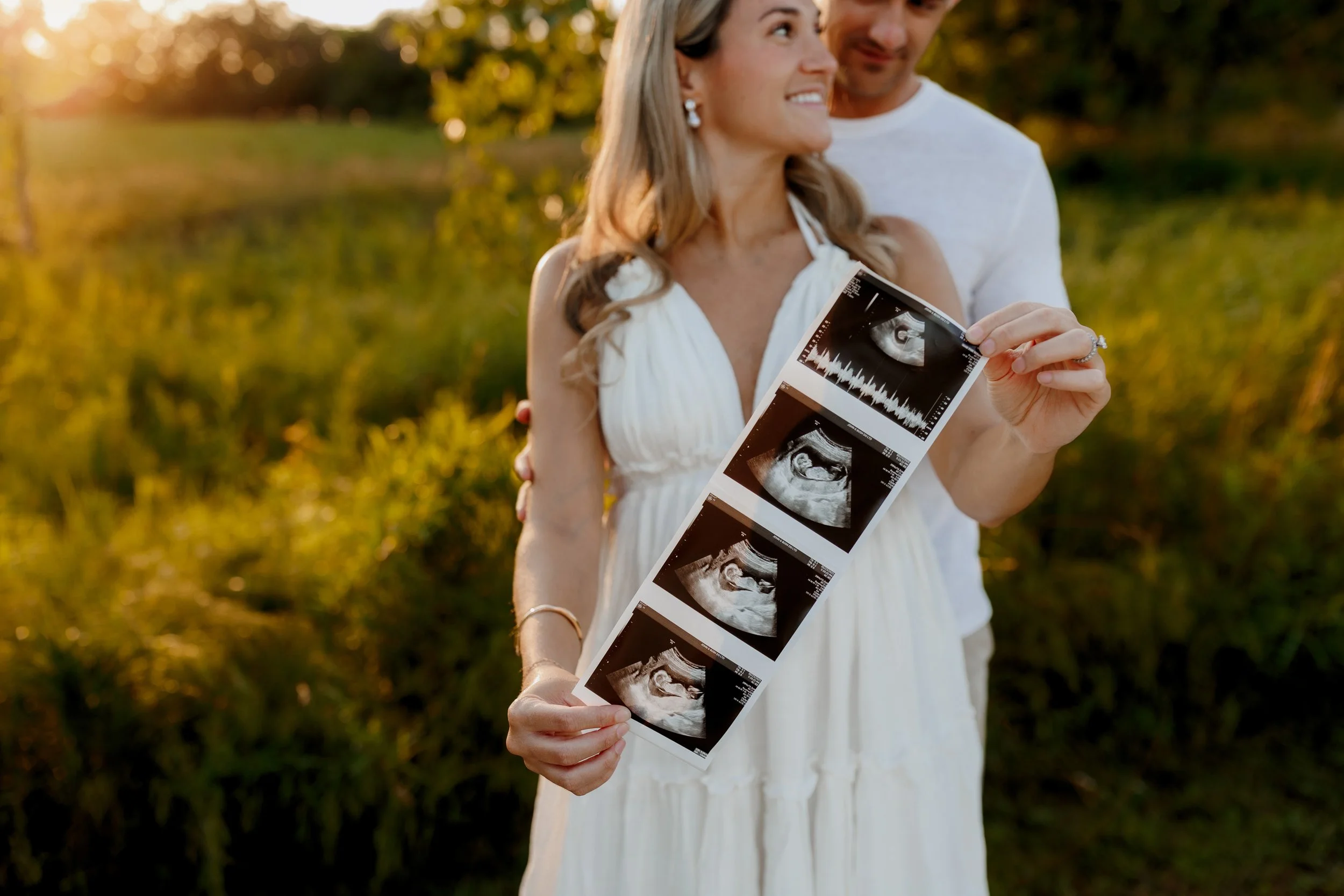 A smiling woman in a white dress holding ultrasound images, standing outdoors in a grassy area during sunset, with a man standing behind her.