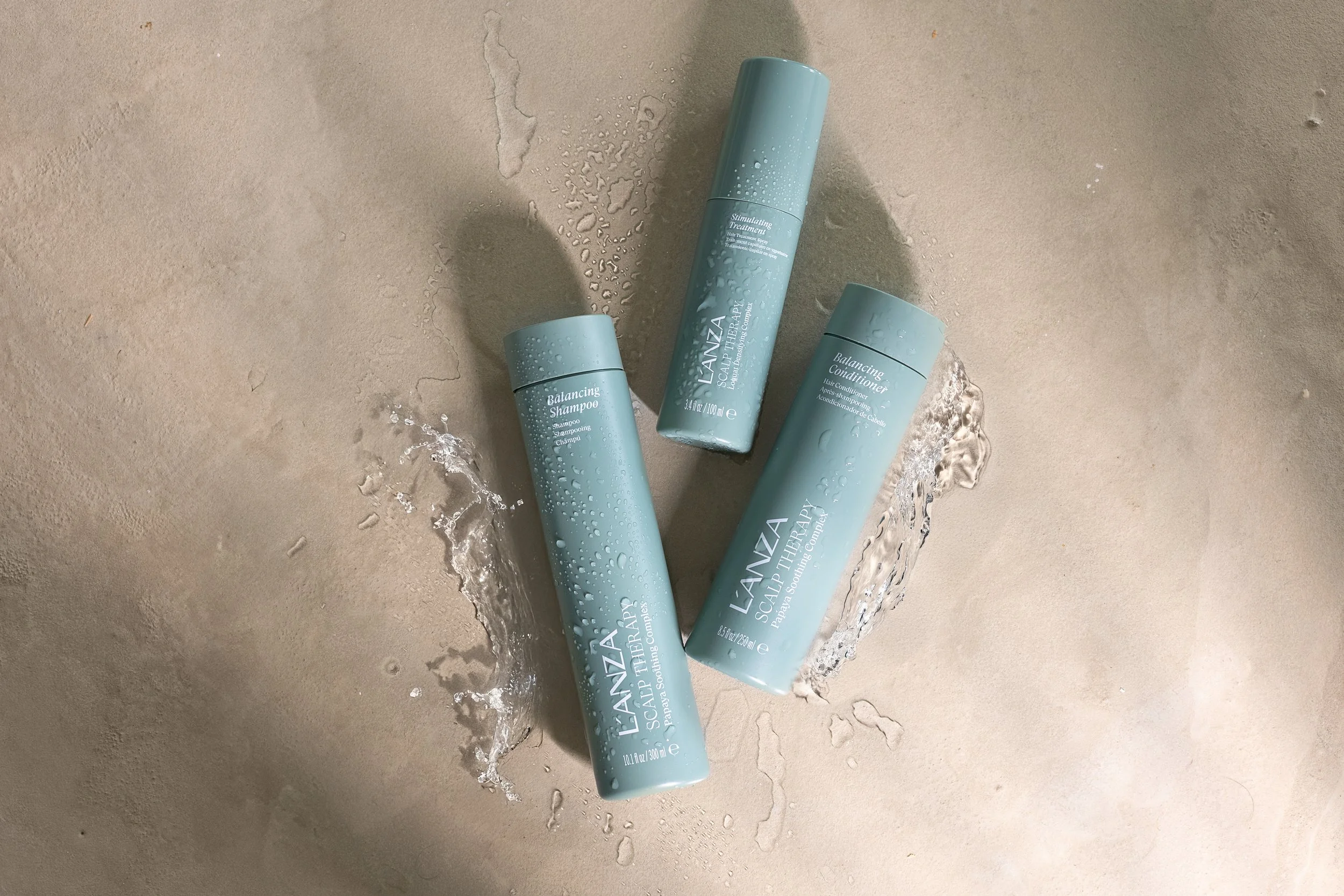 Three L'ANZA scalp therapy hair care bottles on a beige surface with water drops and splashes around them.