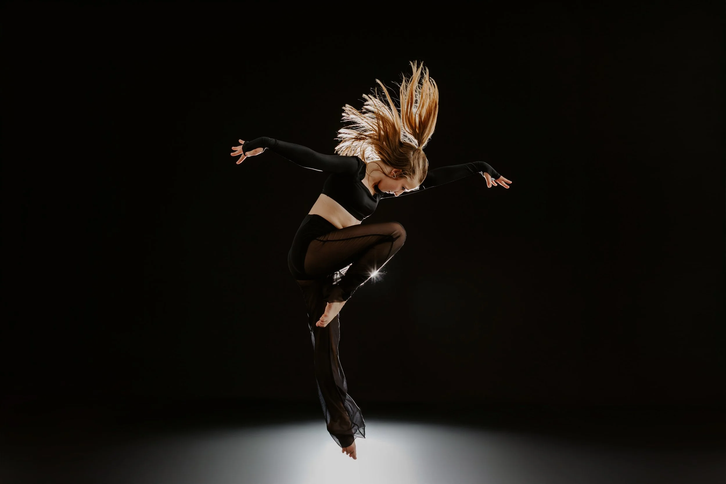A female dancer in black attire with long hair, captured mid-air in a leap against a black background, with lighting creating a spotlight effect.